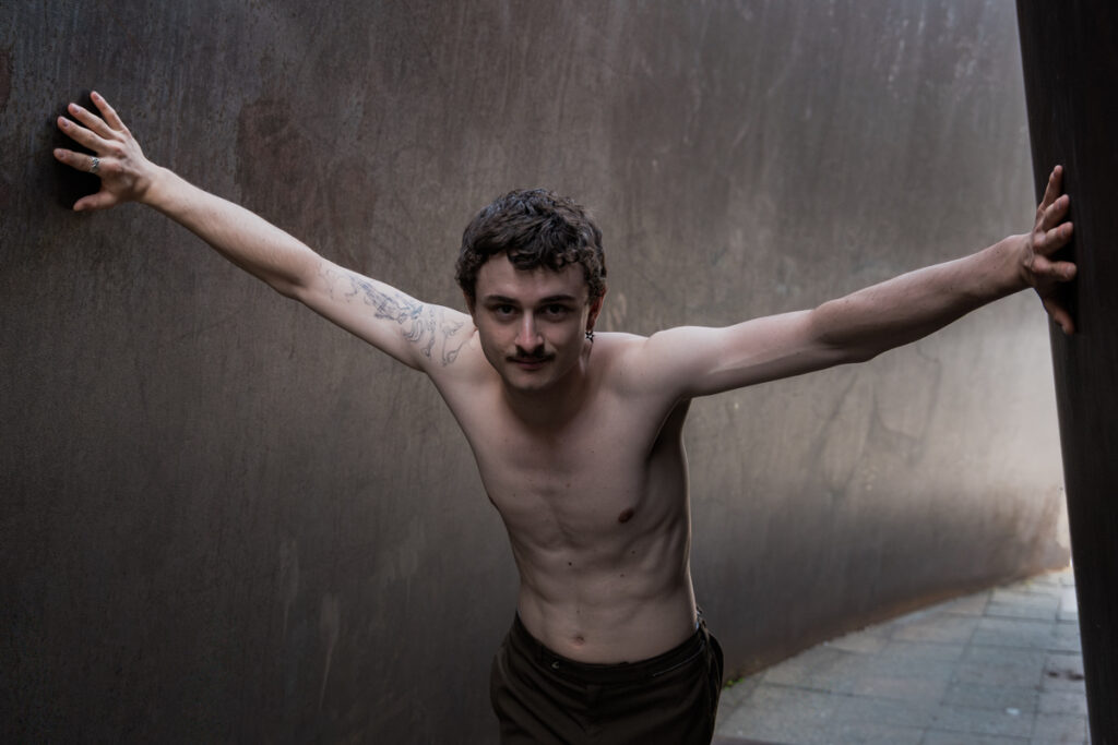 Das Bild zeigt einen Porträt-Szene eines Schauspielers, Felix Steinhilber, der vor einer grauen Metallwand mit ausgestreckten Armen steht. Die Pose vermittelt Kontemplation, Verletzlichkeit und eine gewisse Kraft. Der Hintergrund ist dunkel und unscharf, was die Aufmerksamkeit auf den Schauspieler lenkt.