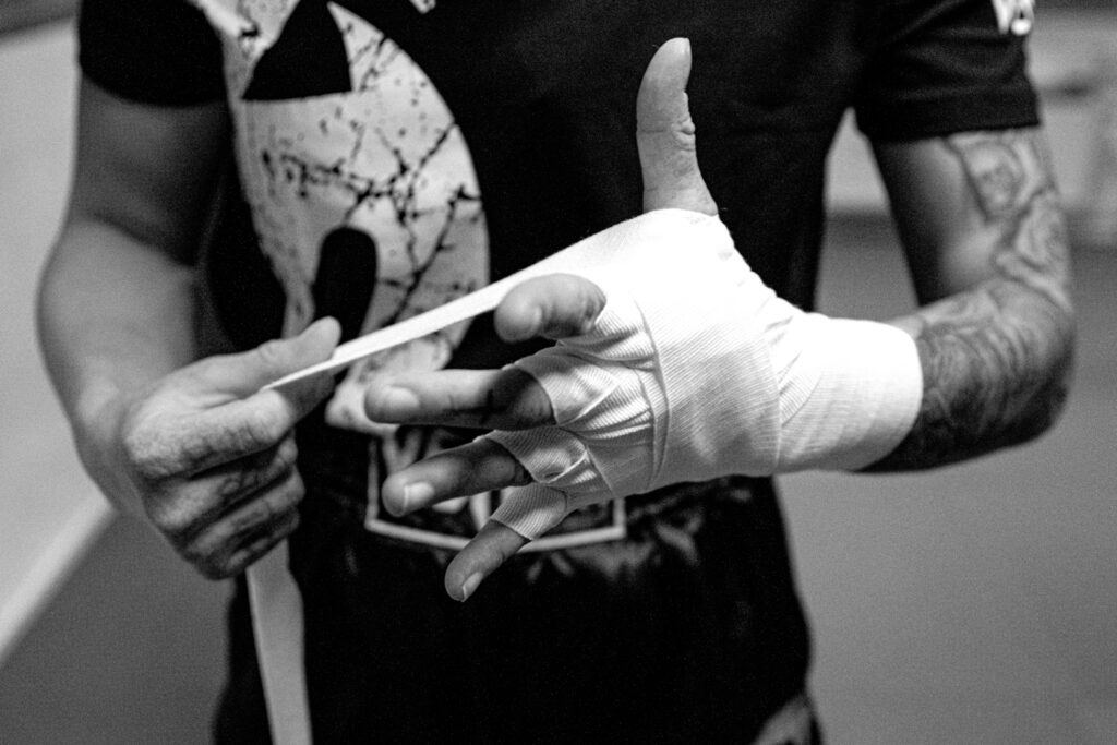Eine Frau wickelt ihre Hand mit einem weißen Kampfbandage ein. Im Hintergrund ist ein dunkler Raum zu sehen.